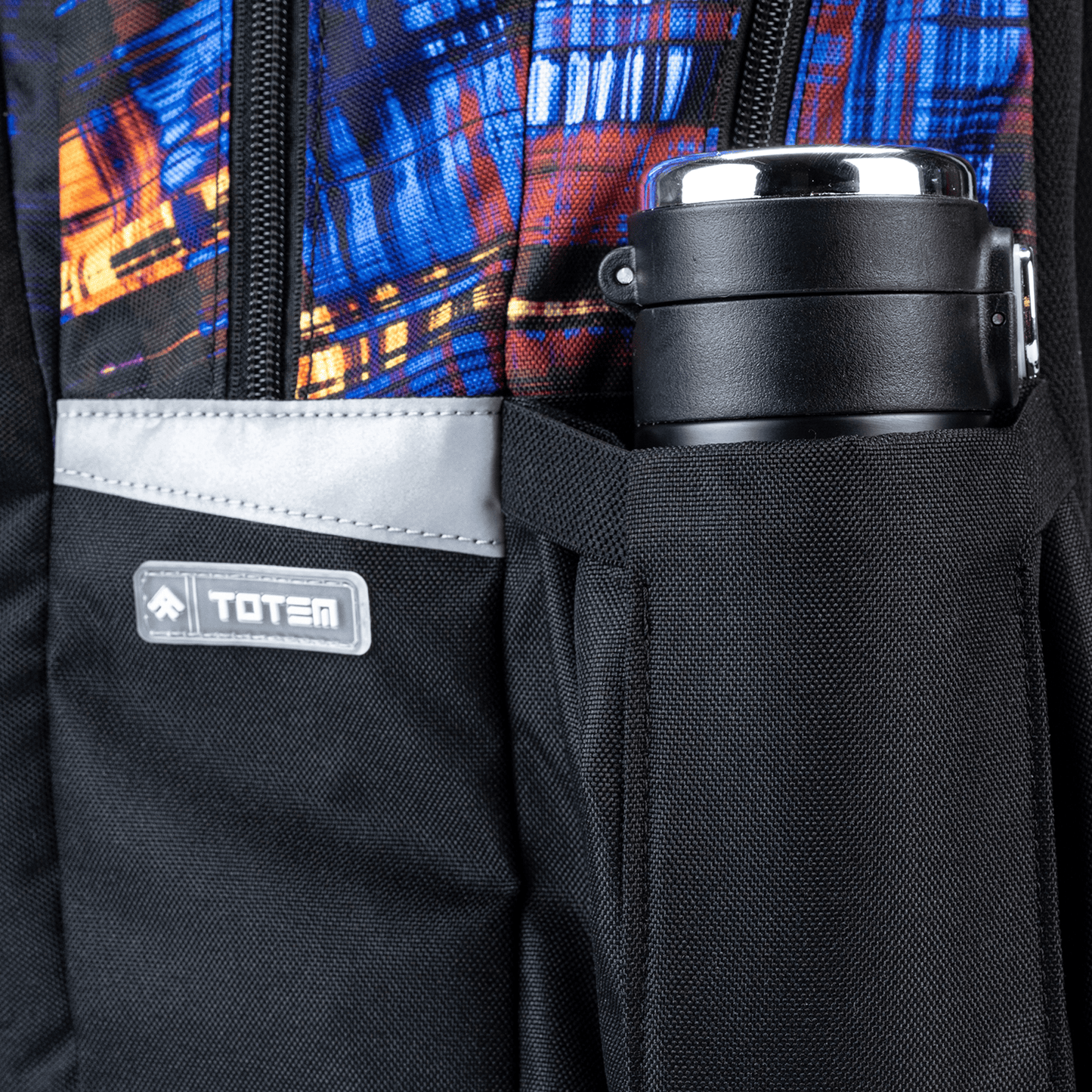 A black side pocket with a stainless flask, grey reflector and blue, orange print, part of a Savvy School bag.