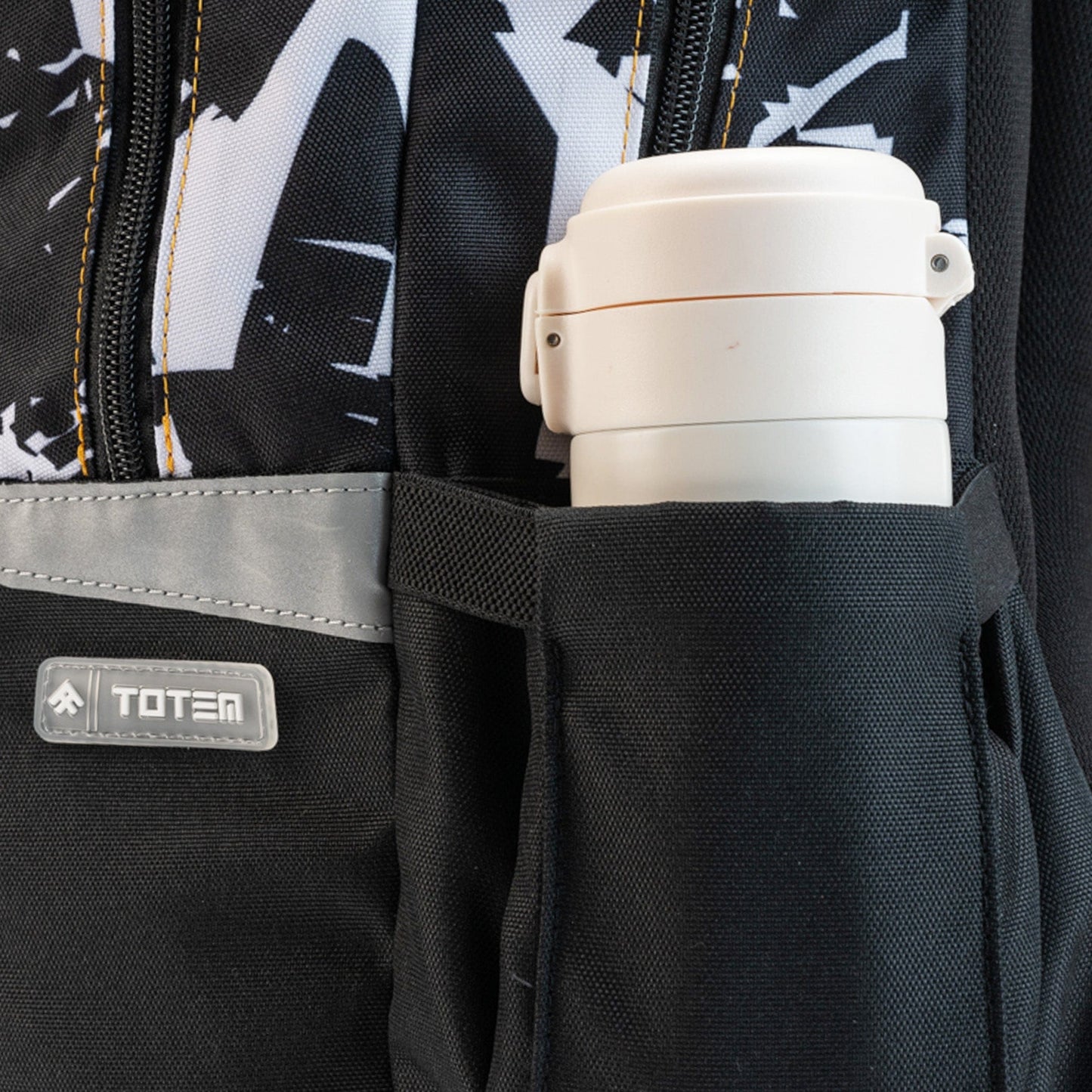 Close-up: black school backpack side pocket with grey reflective trim, Totem badge, and a white bottle.