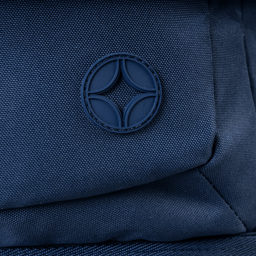 A close-up of a navy Savvy backpack's textured fabric and circular rubber logo.