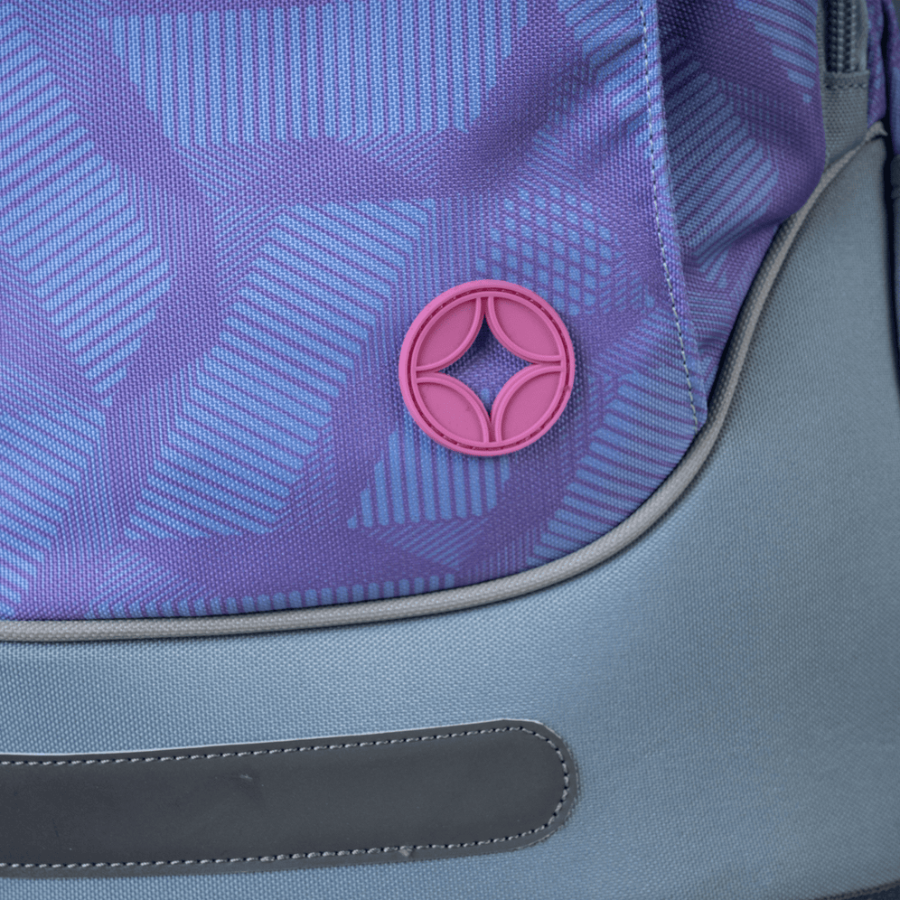 A close-up of a Savvy purple geometric print school backpack featuring a pink circular logo badge and a grey reflective strip.